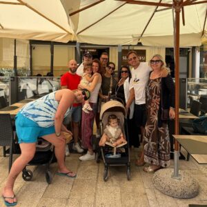 Gruppe von Kolleginnen, Kollegen und Familien mit Kindern und Kinderwagen vor einem Restaurant unter Sonnenschirmen.