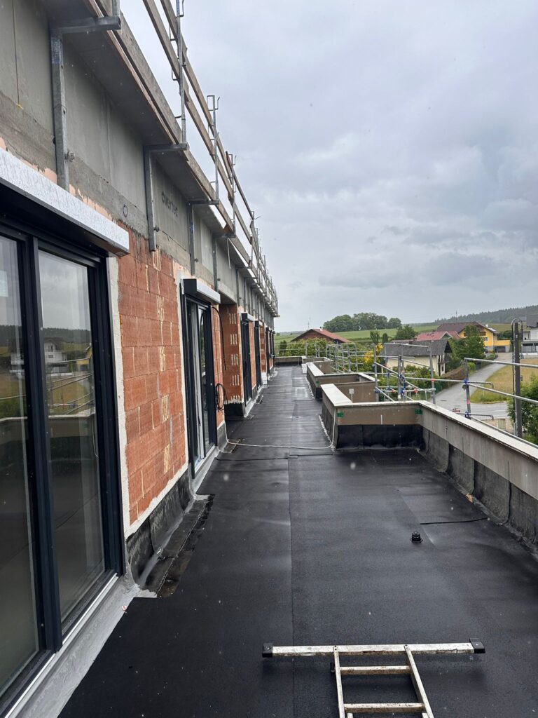 Blick entlang einer noch unvollständig ausgebauten Dachterrasse mit Baugerüst und Blick auf eine Wohngegend.