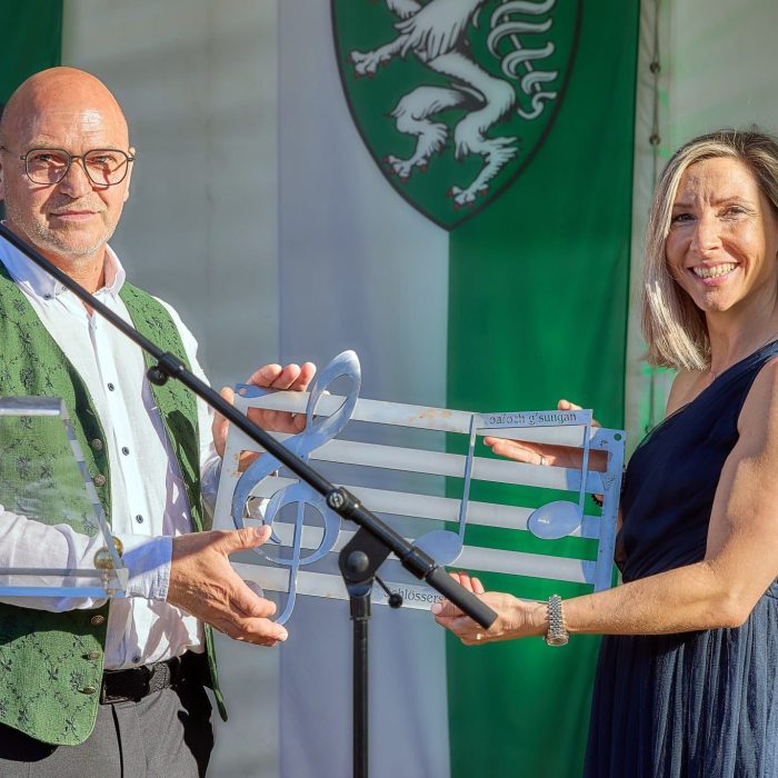 Mann und Frau auf einer Bühne bei der Übergabe eines musikalischen Ehrenpreises vor einer steirischen Flagge.