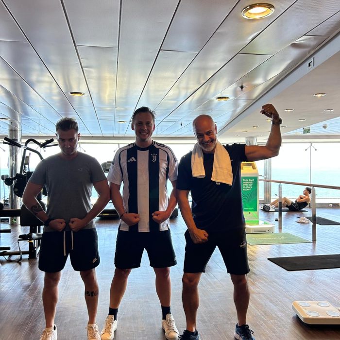 Drei Männer posieren lachend und muskulös im Fitnessbereich eines Kreuzfahrtschiffs.