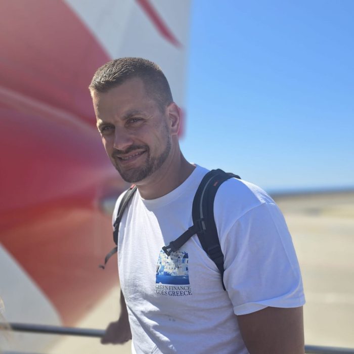 Mann mit Rucksack und ‚Green Finance Goes Greece‘-Shirt steht lächelnd vor einem Flugzeug auf dem Rollfeld.