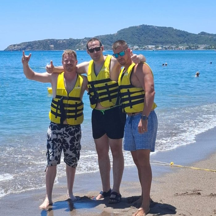 Drei Männer in gelben Rettungswesten stehen am Sandstrand mit Blick auf das Meer und eine grüne Küste.