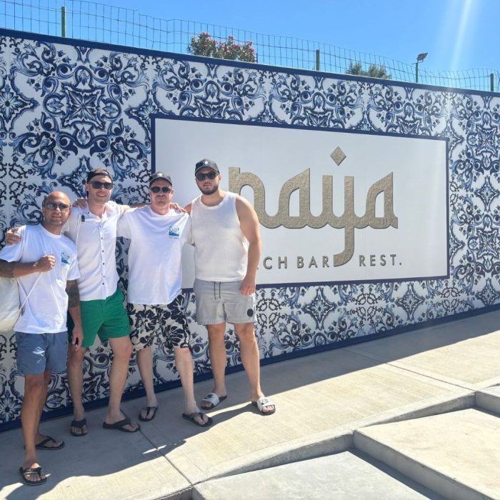 Vier Männer in sommerlicher Kleidung stehen vor einer kunstvoll gefliesten Wand mit ‚Naya Beach Bar Rest.‘-Schriftzug.