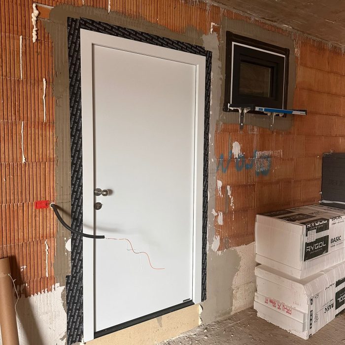 Weiße Eingangstür und kleines Fenster in einem unvollständig verputzten Ziegelmauerwerk mit Baumaterialien daneben.